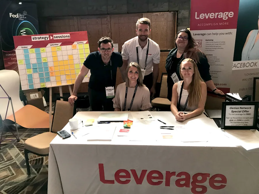 Five of the Leverage team behind their table at the Genius Network annual event: a strategy sessions schedule board covered in sticky notes, a Leverage pull-up banner reading Accomplish More, the Leverage wordmark across the tablecloth, and a Genius Network special offer sign