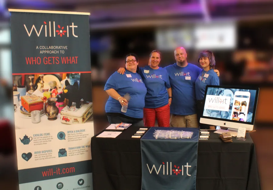 The will-it team of four behind their table at Mass Innovation Nights 102, with a will-it pull-up banner reading a collaborative approach to who gets what, and the product on an iMac